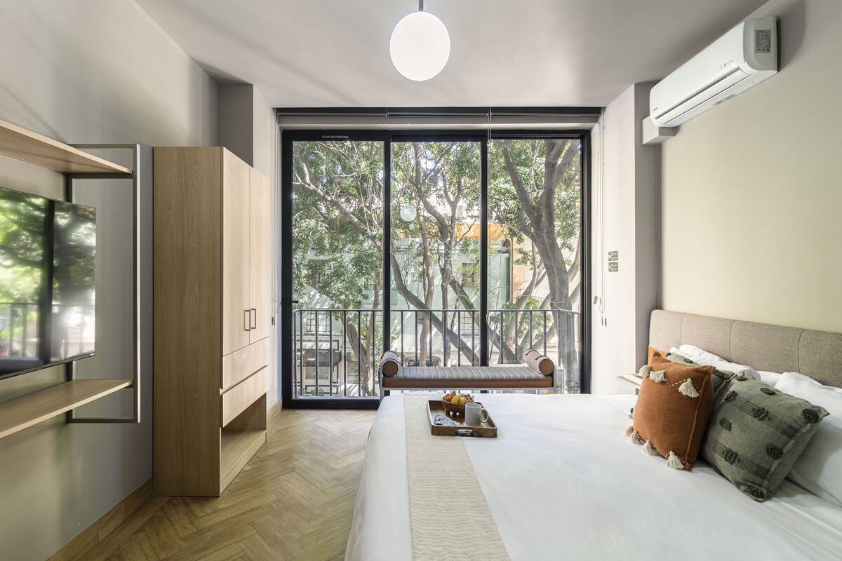 Una habitación de hotel moderna con una cama grande, un televisor, un armario de madera, un balcón con vistas a árboles y un equipo de aire acondicionado.