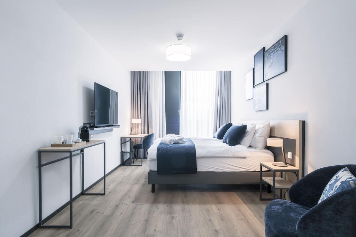 A modern hotel room with a bed, bedside table, chair, desk, and a flat-screen TV.