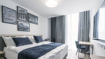 Una habitación de hotel moderna con una cama grande, un escritorio y una silla. Hay cuatro cuadros en la pared y un ventanal con cortinas.