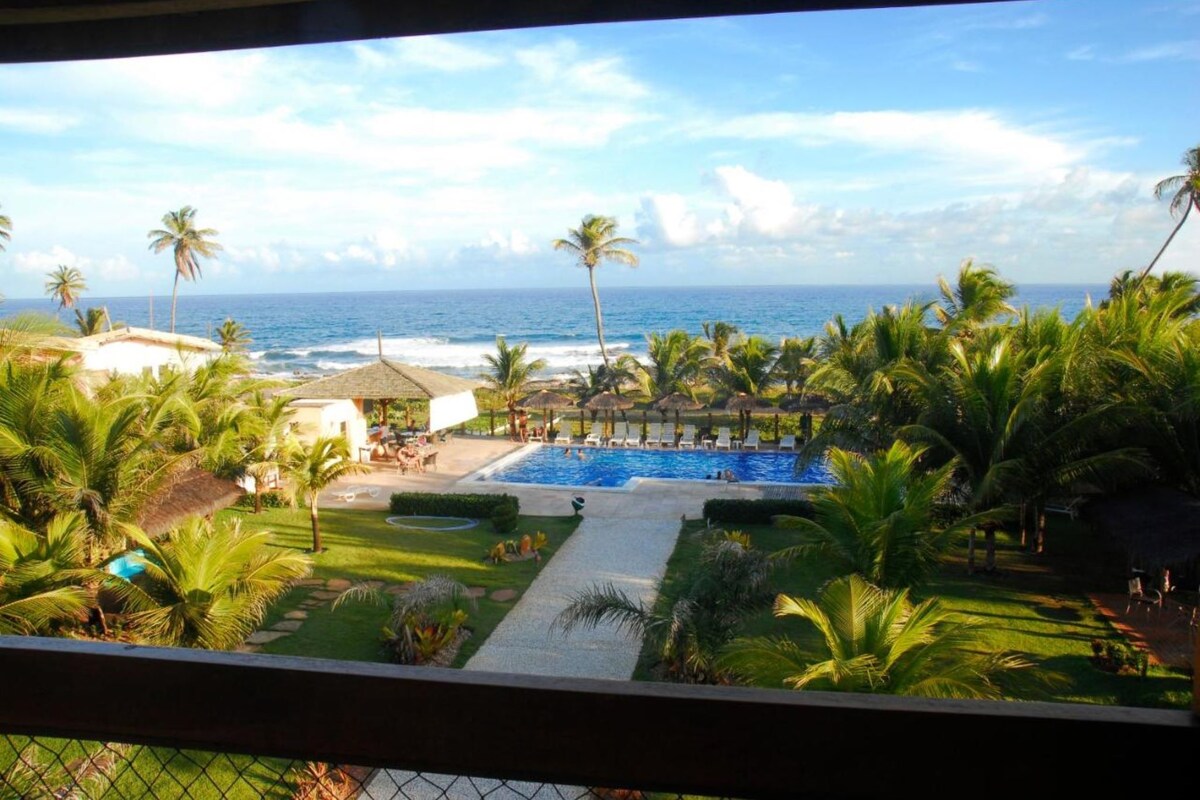 A pool with a view of the ocean, surrounded by palm trees and a thatched-roof structure.