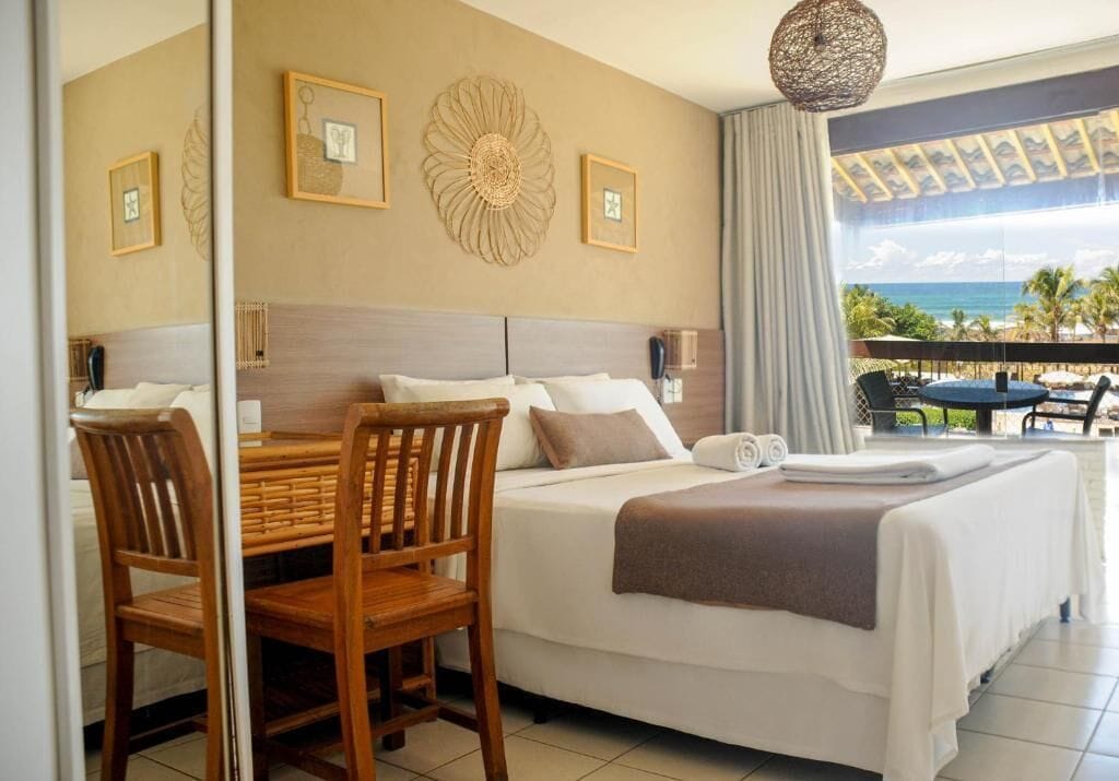 A hotel room with a bed, a dining table with chairs, a balcony with a view of the ocean, and decorative wall art.