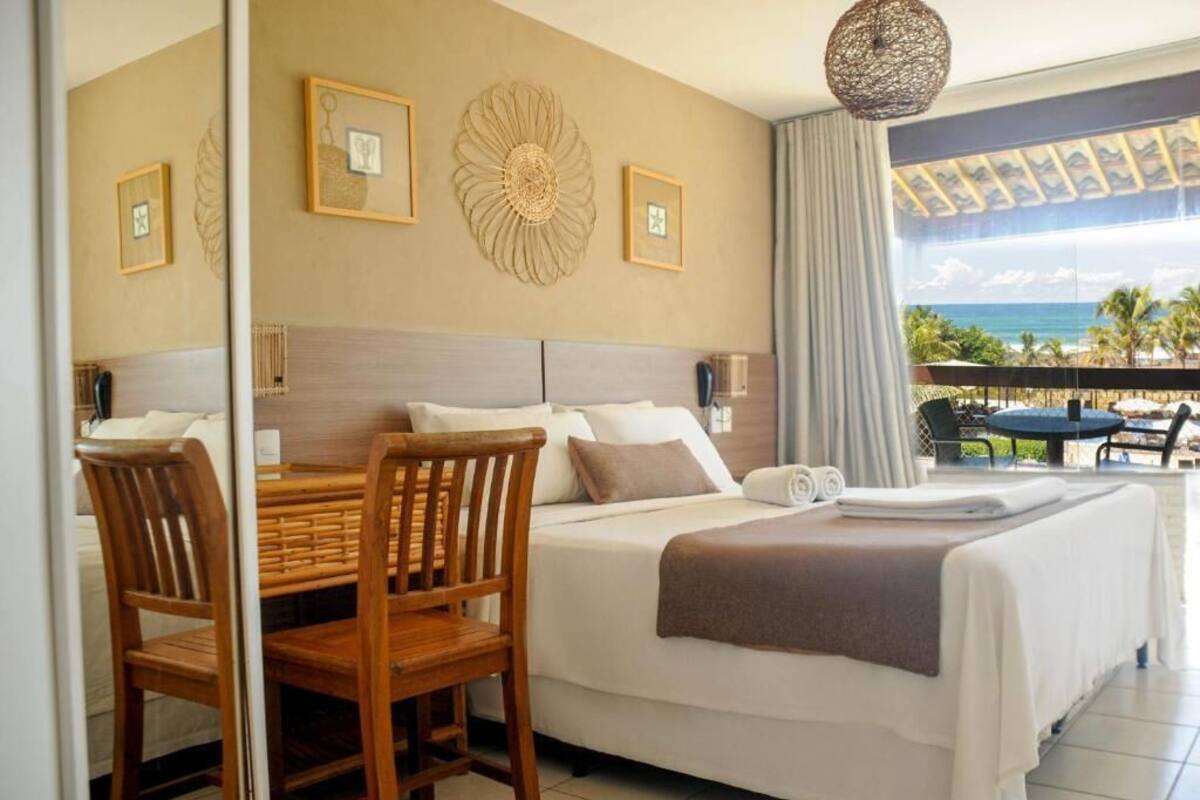 Habitación de hotel con cama, una mesa de comedor con sillas, un balcón con vista al océano y decoración en las paredes.