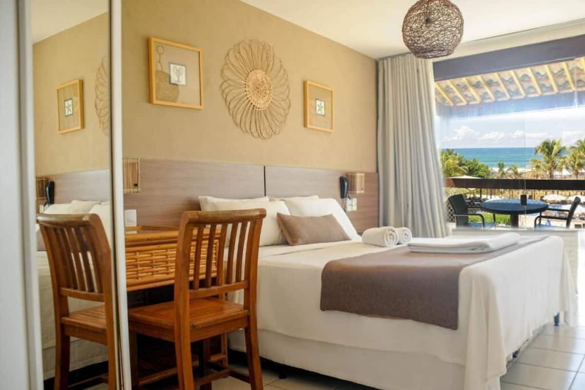A hotel room with a bed, a dining table with chairs, a balcony with a view of the ocean, and decorative wall art.