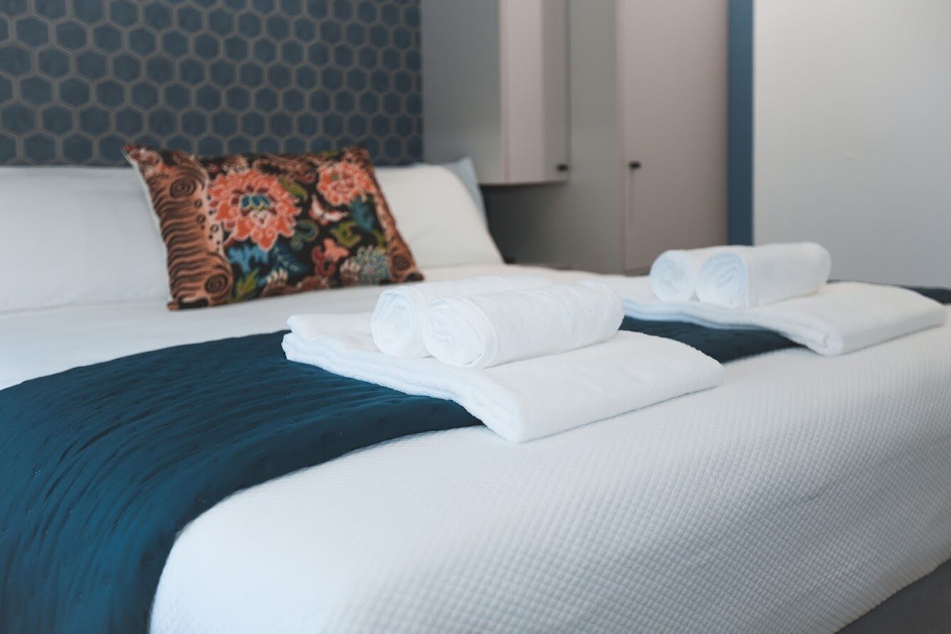 Ein ordentlich bezogenes Bett mit weißen Leinentüchern, einer blauen Decke, einem farbenfrohen Kissen und einem gefalteten Handtuch daneben.
