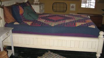A bed with a patterned quilt, a small bedside table, a chair, and a window with a view of trees.