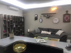 A modern living room with a sofa, a coffee table, a bookshelf, and various clocks on the wall. - Stunning Vacation Rental in Dbayeh, at a Prime Location, Between Le Mall and Abc (Dbayeh)