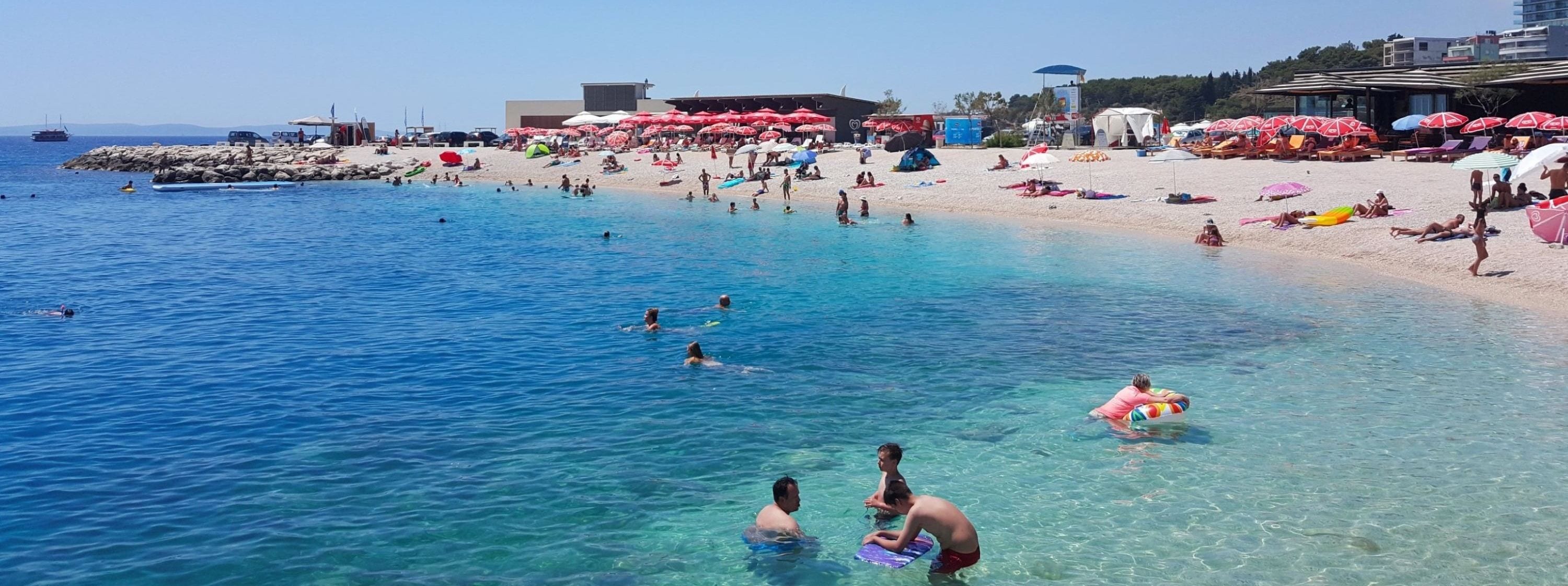 Une plage avec une eau claire, des gens qui se baignent et prennent des bains de soleil, et des parasols.