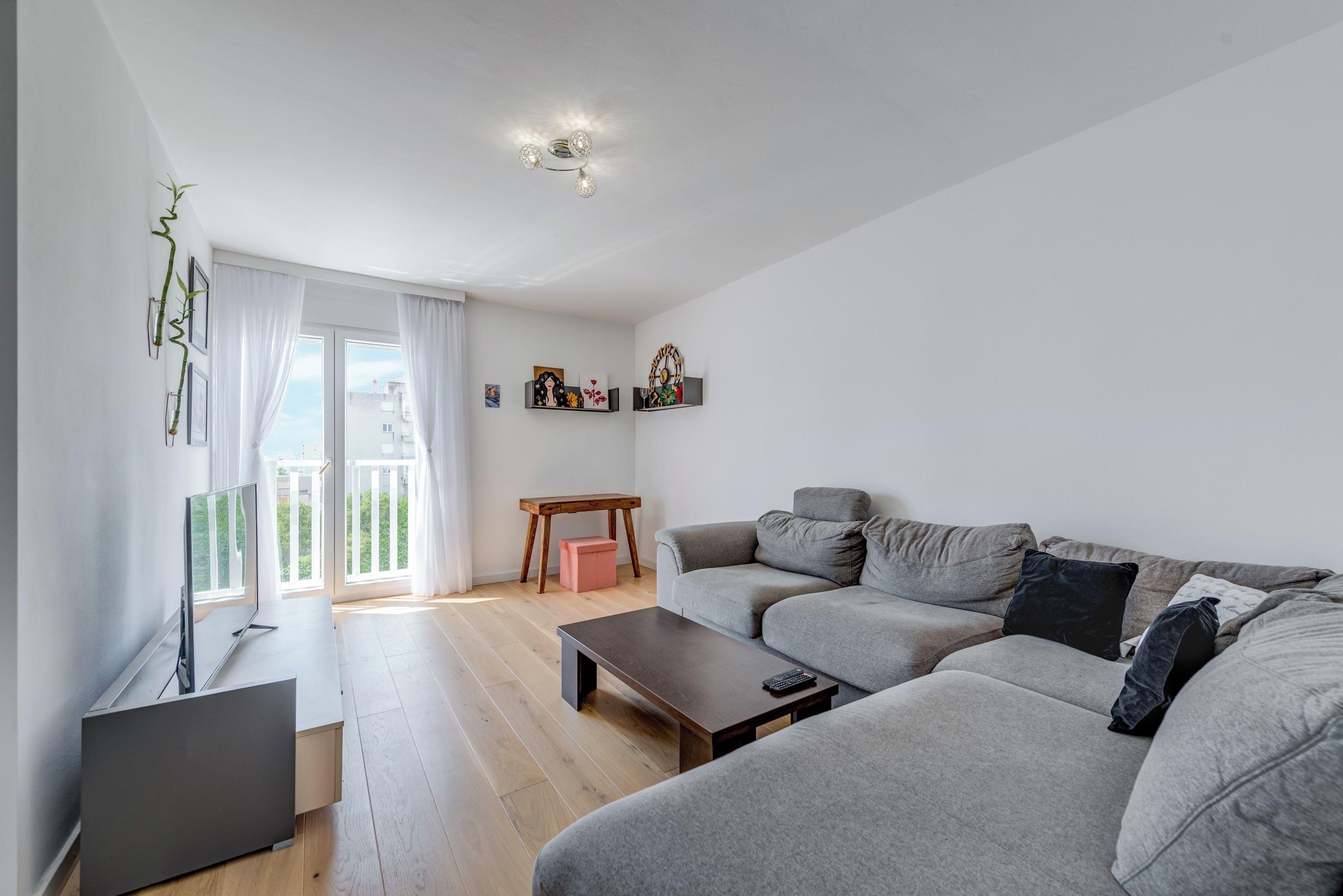 A modern living room with a grey sofa, a coffee table, and a TV on a stand. There is a window with white curtains, a small table, and a shelf with decorative items.