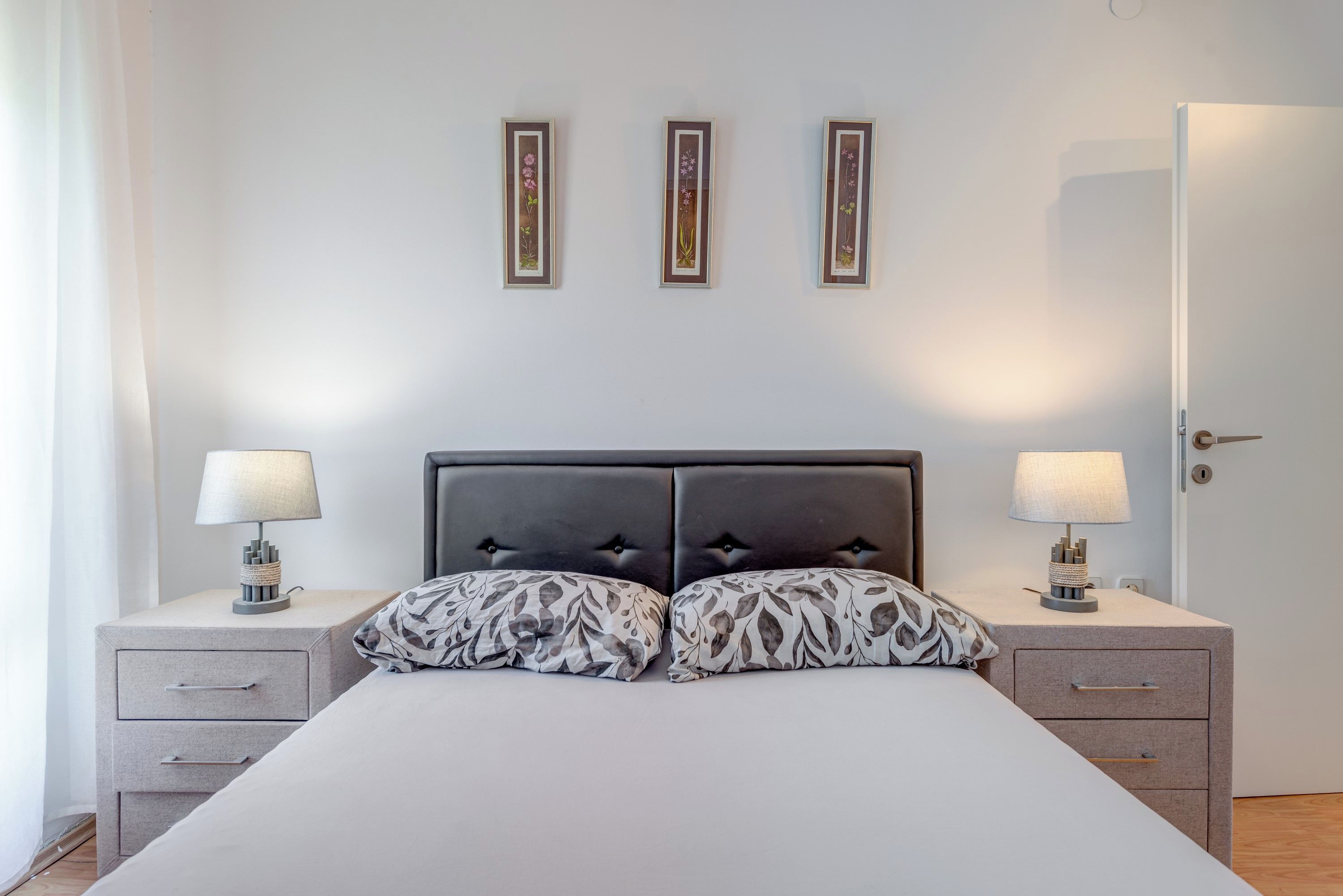 Quarto com cama, duas mesas de cabeceira com luminárias, uma porta e três quadros na parede.