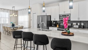 A modern kitchen with white cabinets, a central island with bar stools, a refrigerator, and a coffee machine. - Embark on a Magical Family Getaway:Themed bd, PV Pool, PS5, Air Hockey, Resort (Kissimmee)