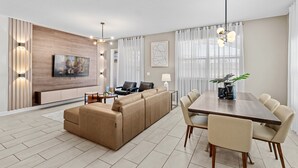 A modern living room with a sofa, dining table, and chairs, a TV mounted on the wall, and a large window with curtains. - Embark on a Magical Family Getaway:Themed bd, PV Pool, PS5, Air Hockey, Resort (Kissimmee)