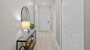A hallway with a white door at the end, a console table with a lamp and a plant, and a tiled floor. - Embark on a Magical Family Getaway:Themed bd, PV Pool, PS5, Air Hockey, Resort (Kissimmee)
