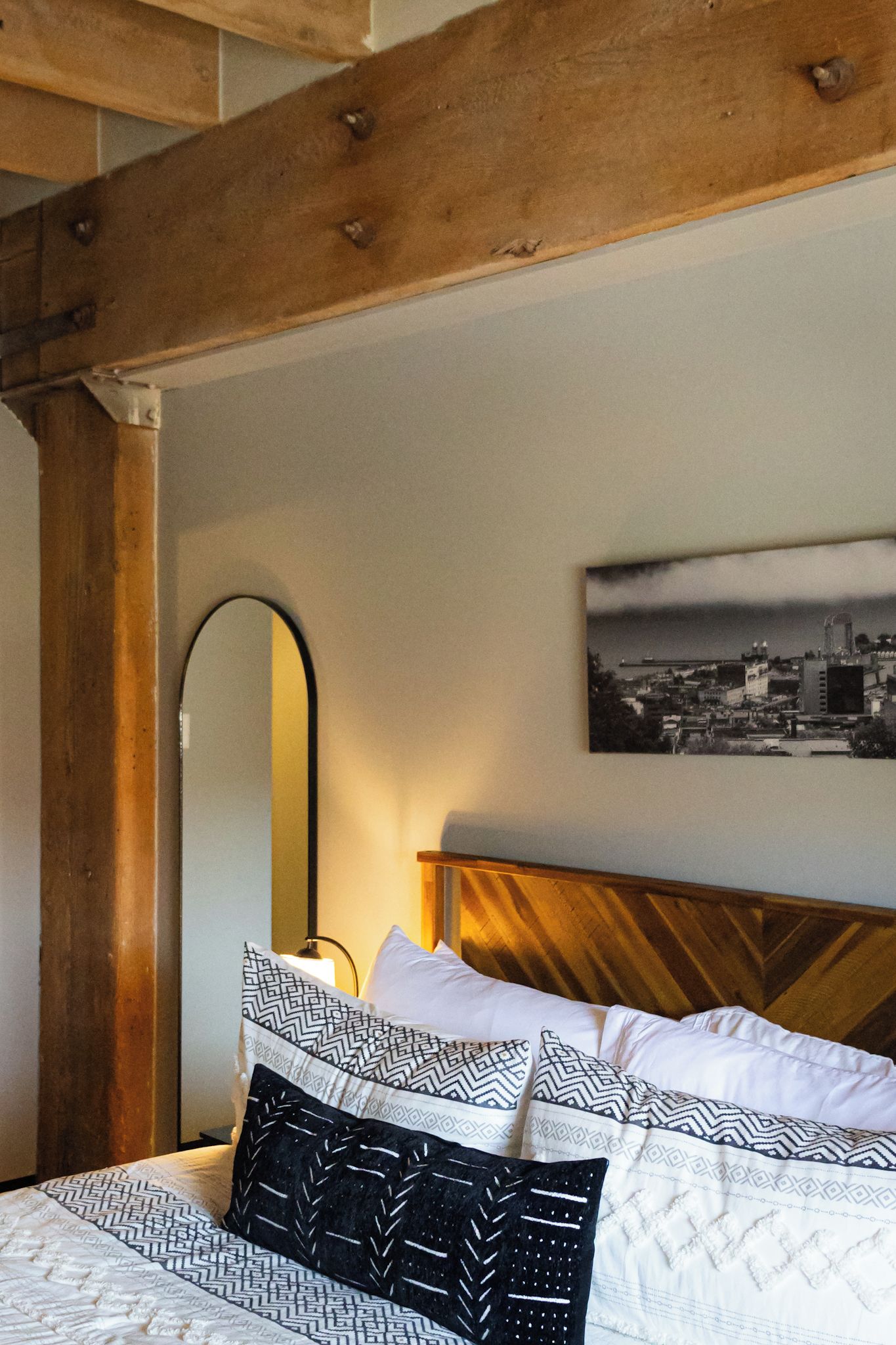 Een bed met gestreepte kussens, een houten hoofdbord, een stadsgezicht schilderij en een spiegel aan de muur.