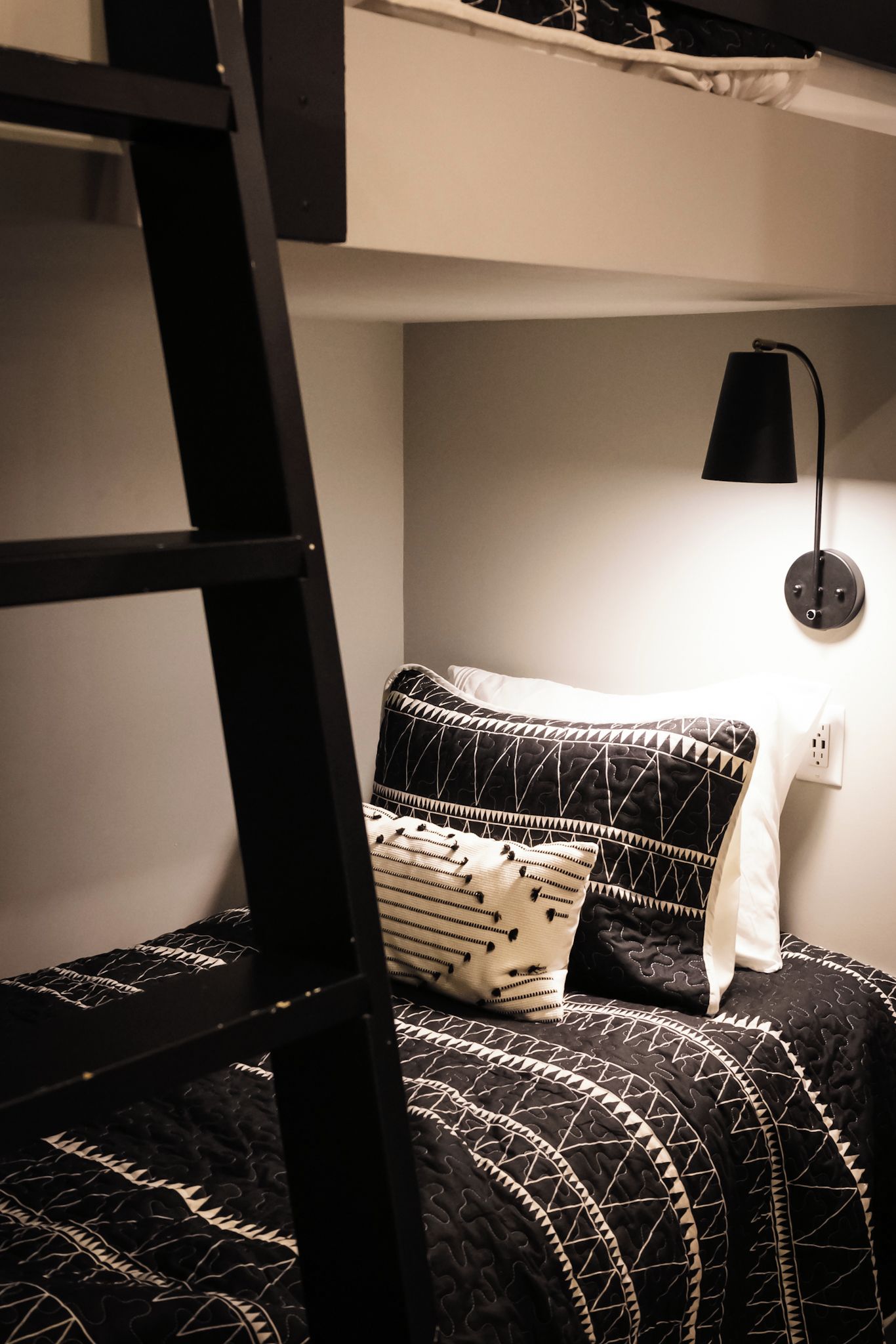 Ein Stockbett mit schwarzer Leiter, einer gemusterten schwarz-weißen Decke und einer einzelnen schwarzen Wandlampe.