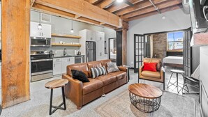 A spacious living area with a brown leather sofa, a wooden coffee table, and a leather armchair. A kitchen with modern appliances is visible in the background. - Aurora Black | The Brix | Pool in Canal Park! (Duluth)