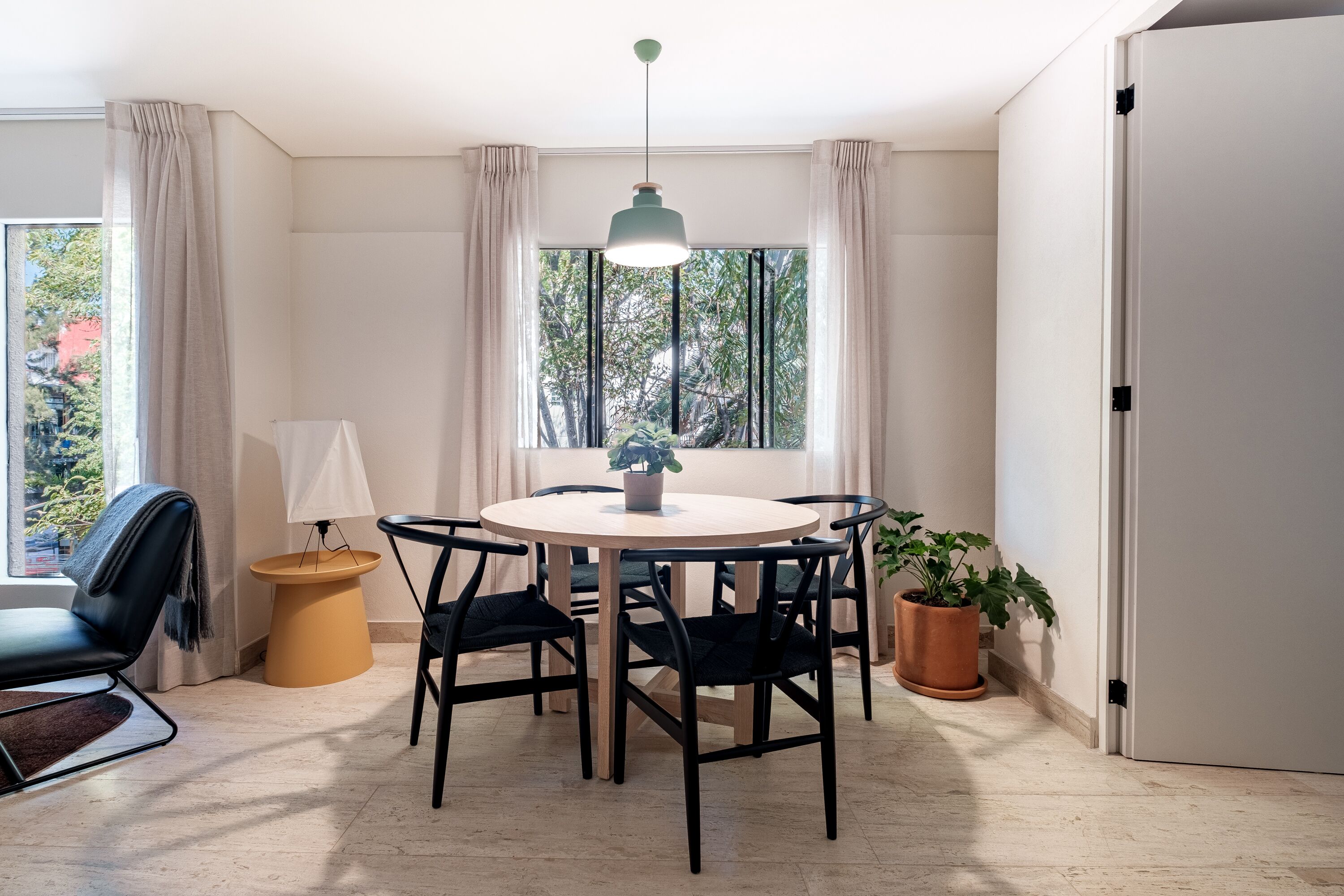 A dining area with a round table, black chairs, a hanging light fixture, a potted plant, and a view of trees through the window.