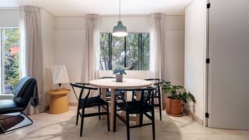 A dining area with a round table, black chairs, a hanging light fixture, a potted plant, and a view of trees through the window.