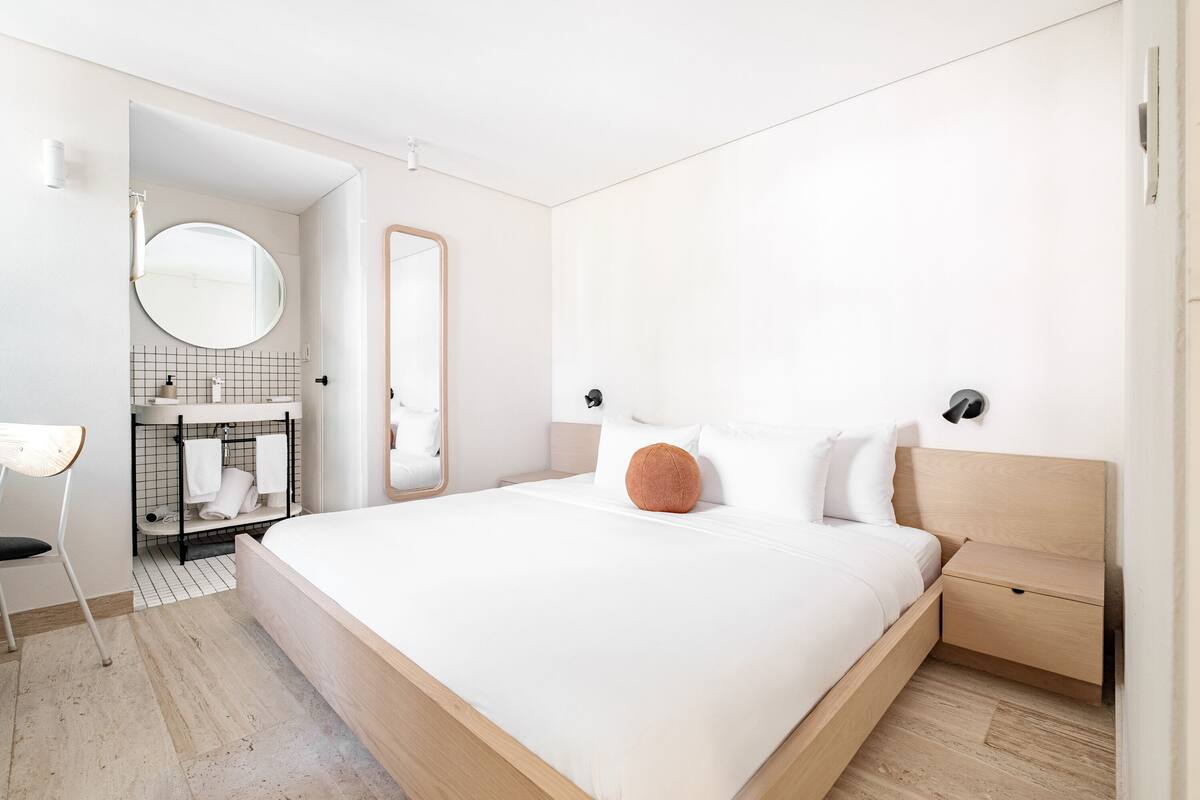 Ein modernes Schlafzimmer mit einem großen Bett, einem Nachttisch, einem Spiegel und einem Badezimmer mit Waschbecken und Handtuchhalter.