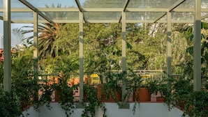 Una estructura de invernadero con paredes de vidrio y marcos de metal, rodeada de abundante vegetación y plantas.