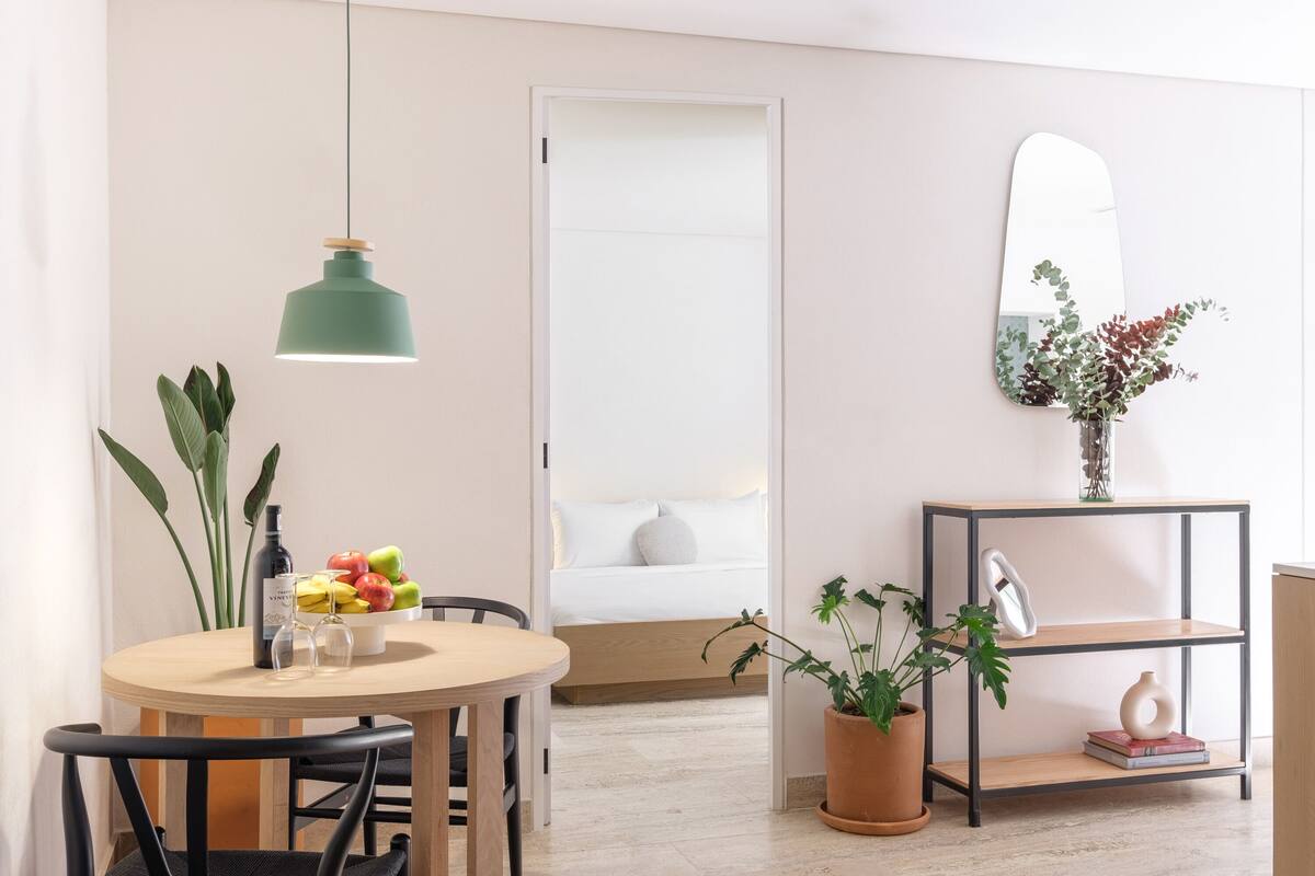 Ein modernes Zimmer mit einem runden Holztisch, einer grünen Pendelleuchte, einem Blumentopf, einem Spiegel und einem Bett mit weißer Bettwäsche.