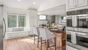 A kitchen with white cabinetry, a built-in bench with chairs, a large window, and a wall-mounted TV. - Hot Tub EV Charger Pool Dog OK Grill Tennis Hiking (Wintergreen Resort)