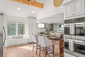 A kitchen with white cabinetry, a built-in bench with chairs, a large window, and a wall-mounted TV.
