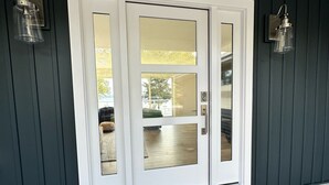 A dark blue exterior wall with a white door featuring a glass panel. There are two wall-mounted lanterns on either side of the door. A brown doormat with "WELCOME" is placed in front of the door. - Waterfront Lakehouse - Completely Remodeled - Watersports + Hot Tub (Elk Rapids)