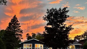 A two-story house with a balcony, outdoor seating, and a large tree in front. - Waterfront Lakehouse - Completely Remodeled - Watersports + Hot Tub (Elk Rapids)