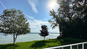 A deck with white chairs overlooking a lake, surrounded by trees and a clear sky. - Waterfront Lakehouse - Completely Remodeled - Watersports + Hot Tub (Elk Rapids)