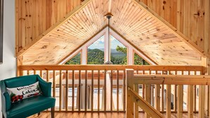 A cabin with a wooden interior, a balcony with a view of the forest, and a green chair with a welcome pillow. - Black Bear Cabin w Majestic Views (Fairview)