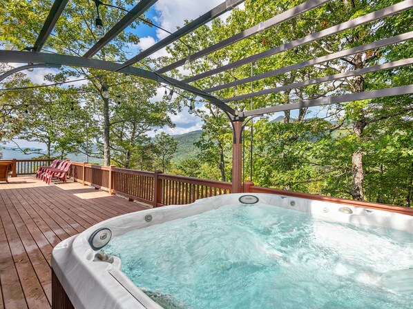 A hot tub on a wooden deck with a pergola overhead, surrounded by trees and a view of a mountainous landscape.