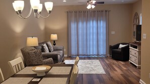 A dining area with a wooden table, chairs, and a chandelier. There is a sofa with cushions and a television on a stand in the background.