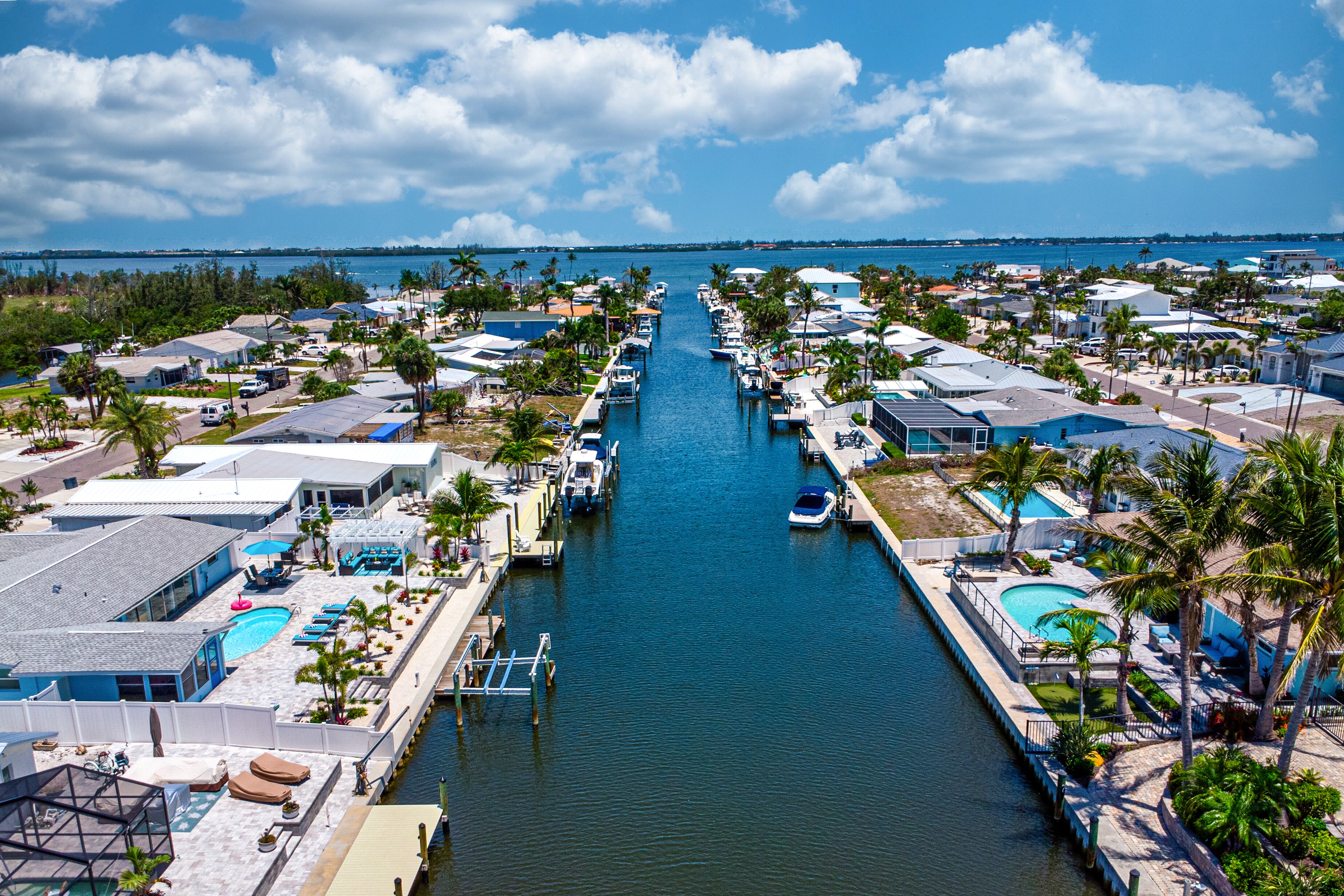 A marina with boats docked, a waterway, and a row of houses with pools and palm trees.