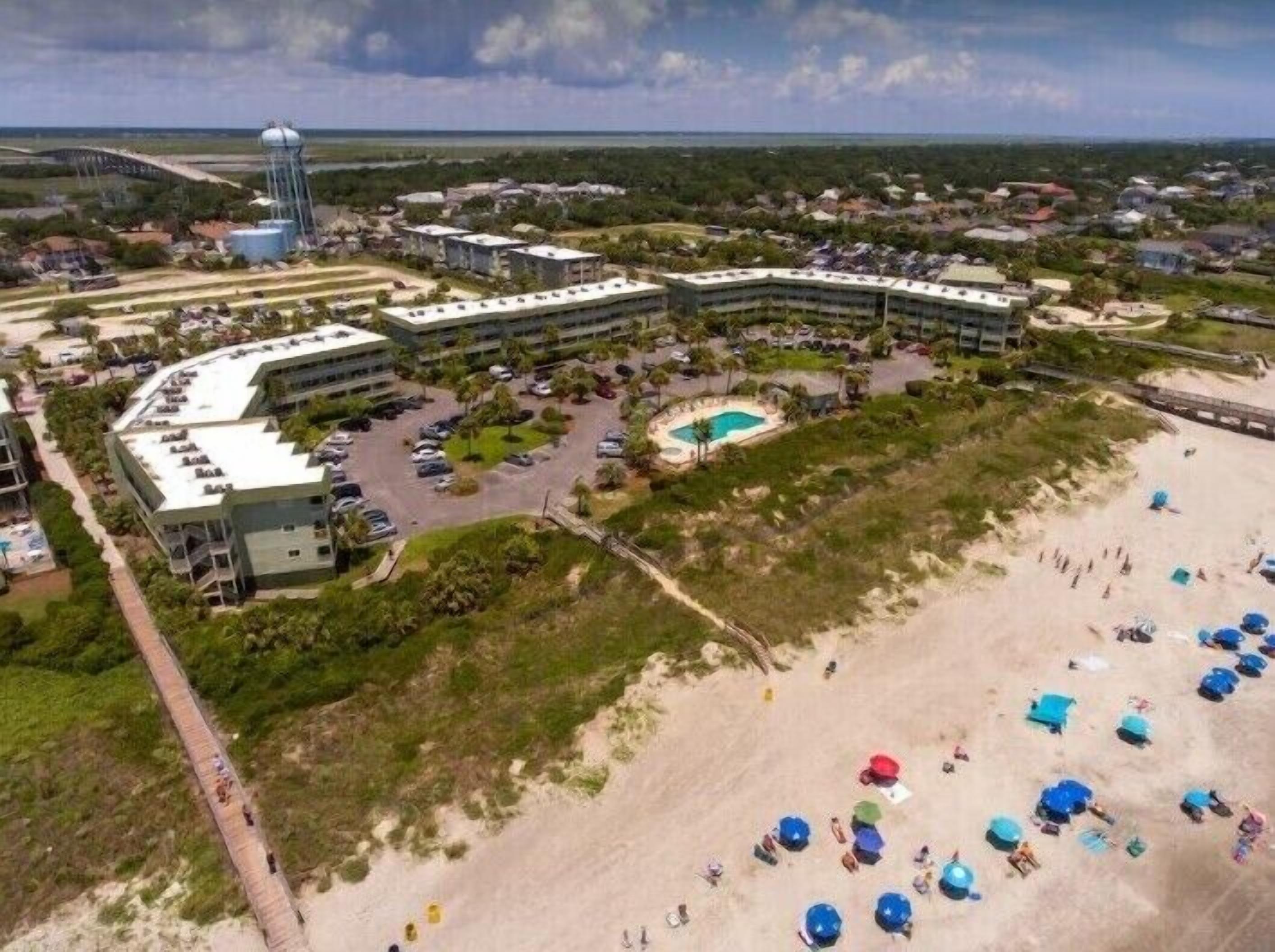 An aerial view of a coastal resort with multiple buildings, a swimming pool, and a beach area with umbrellas.