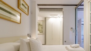Habitación de hotel con cama, almohadas, una mesita de noche, un cuadro enmarcado y una puerta que da a otra habitación.