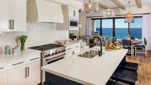 Een moderne keuken met witte kasten, een groot eiland met een spoelbak, en uitzicht op de oceaan door grote ramen.