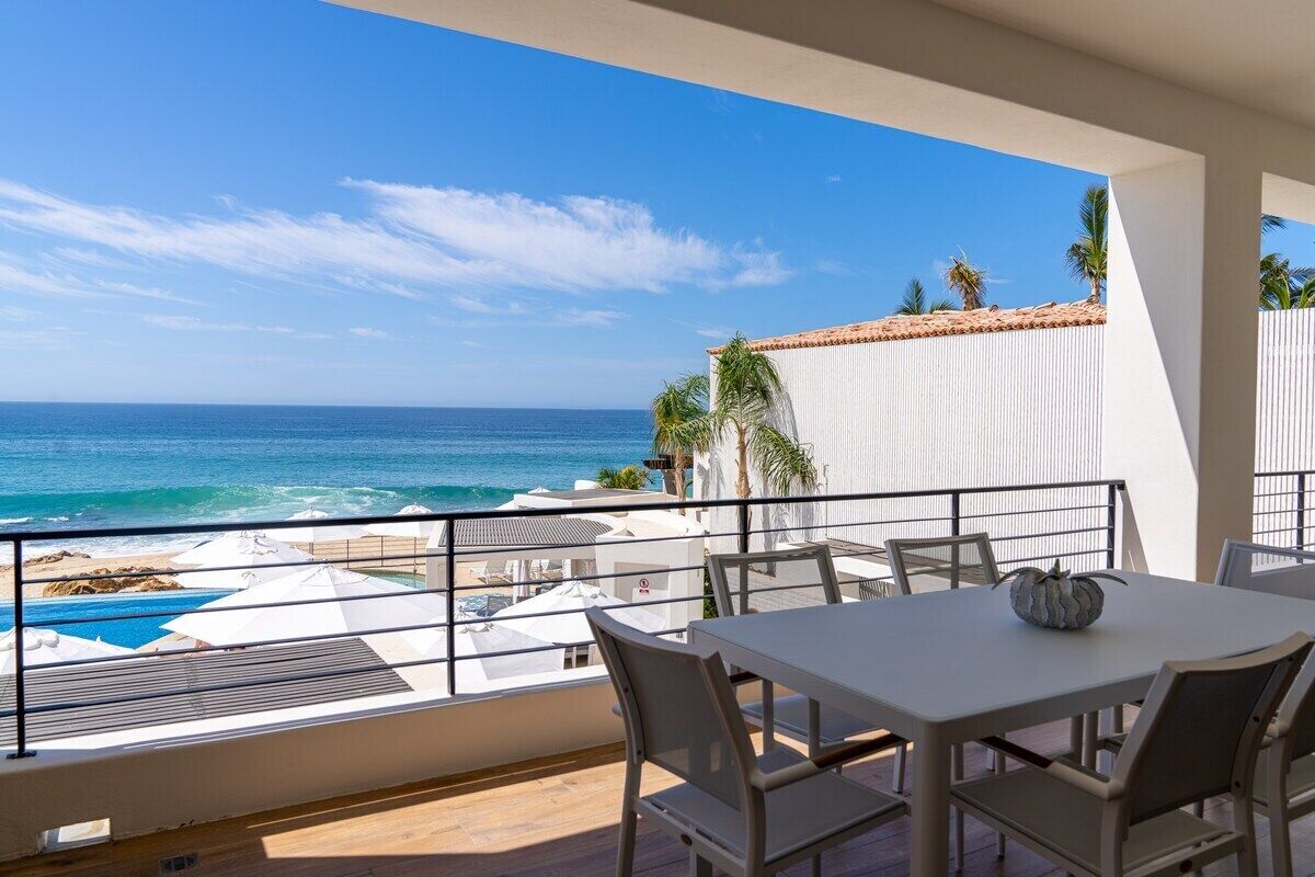 A balcony with a view of the ocean, a pool, and beach umbrellas.