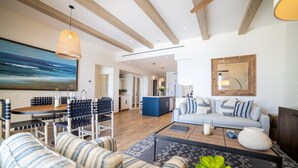 Een moderne woonkamer met een bank, eettafel en stoelen, een keuken op de achtergrond, en een groot schilderij aan de muur.