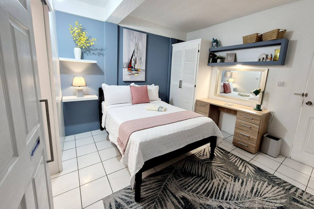 A bedroom with a bed, bedside table, lamp, dresser, mirror, and a wall with a painting and shelf.