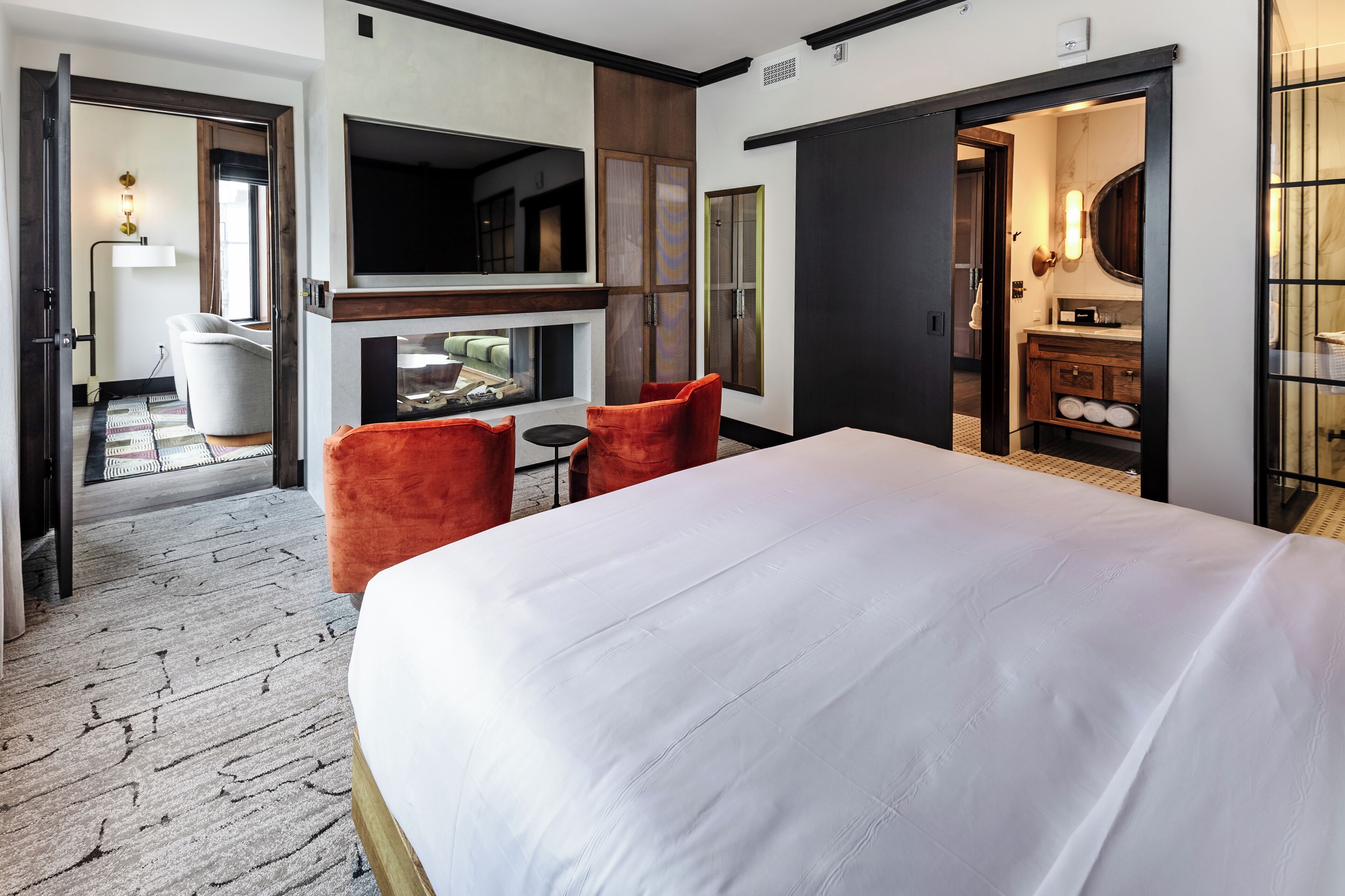 A hotel room with a large bed, two red chairs, a TV, a fireplace, and a bathroom visible through an open door.