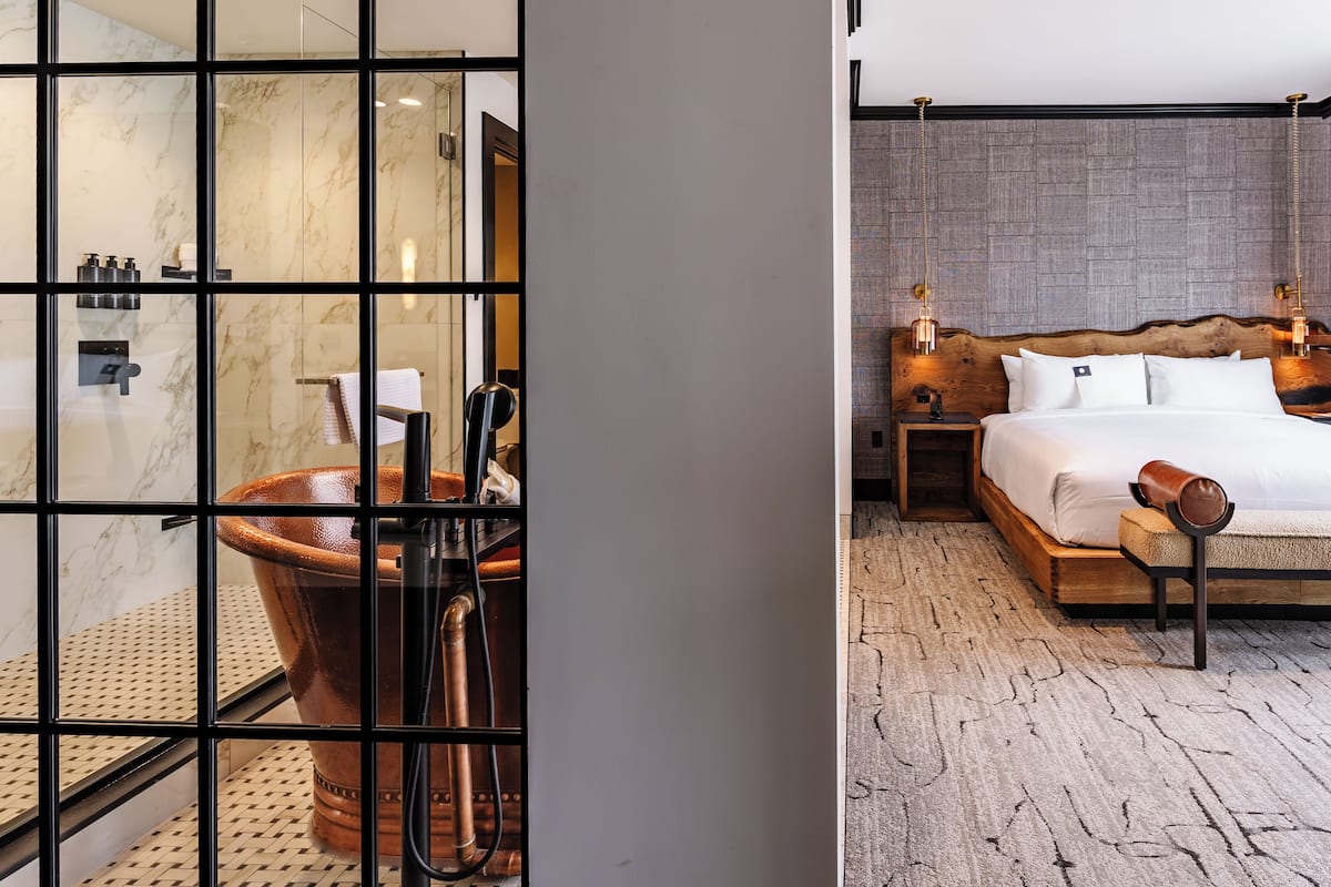 A modern bathroom with a glass-enclosed shower, a large terracotta bathtub, and a marble wall. The bedroom features a wooden bed frame, a large bed, and a textured rug.