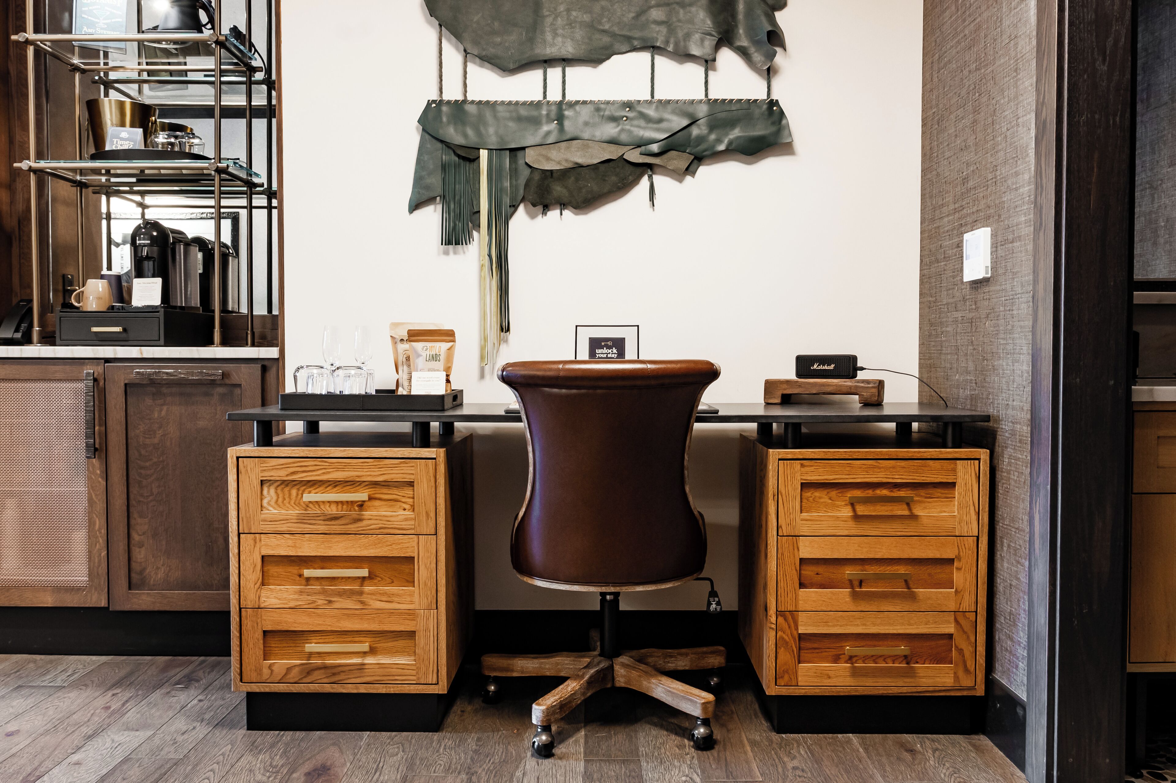 A modern office space with a desk, two wooden drawers, a brown chair, a shelf with kitchen items, and a wall-mounted decoration.