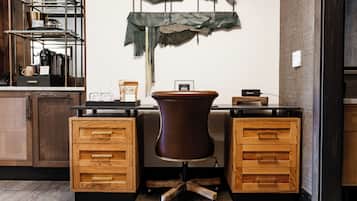 A modern office space with a desk, two wooden drawers, a brown chair, a shelf with kitchen items, and a wall-mounted decoration.