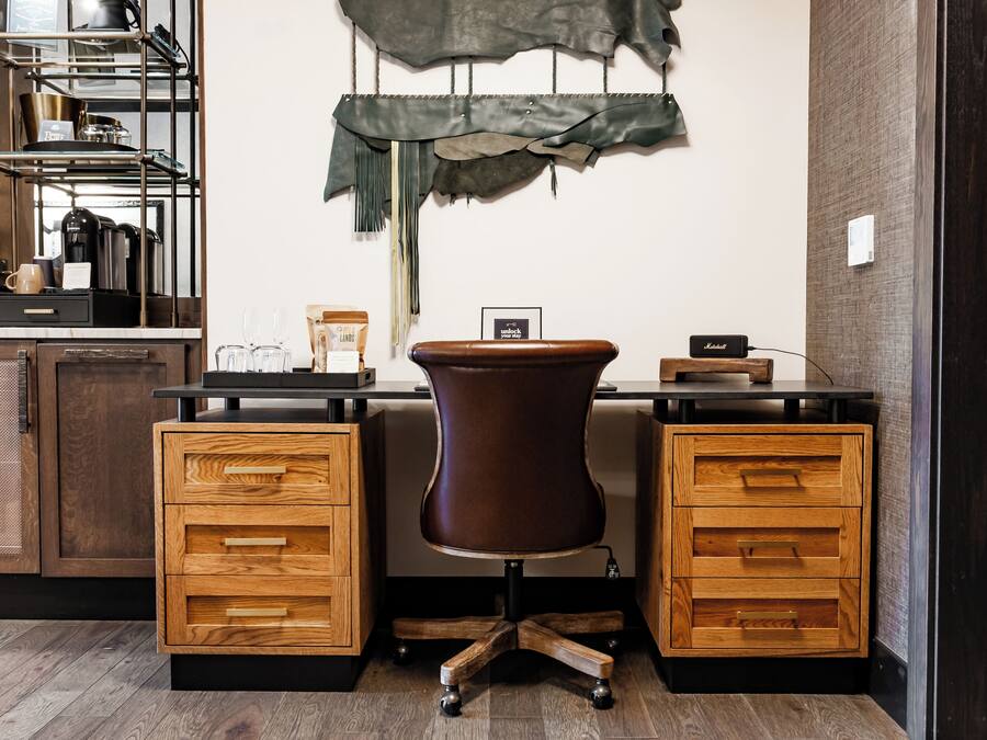 A modern office space with a desk, two wooden drawers, a brown chair, a shelf with kitchen items, and a wall-mounted decoration.