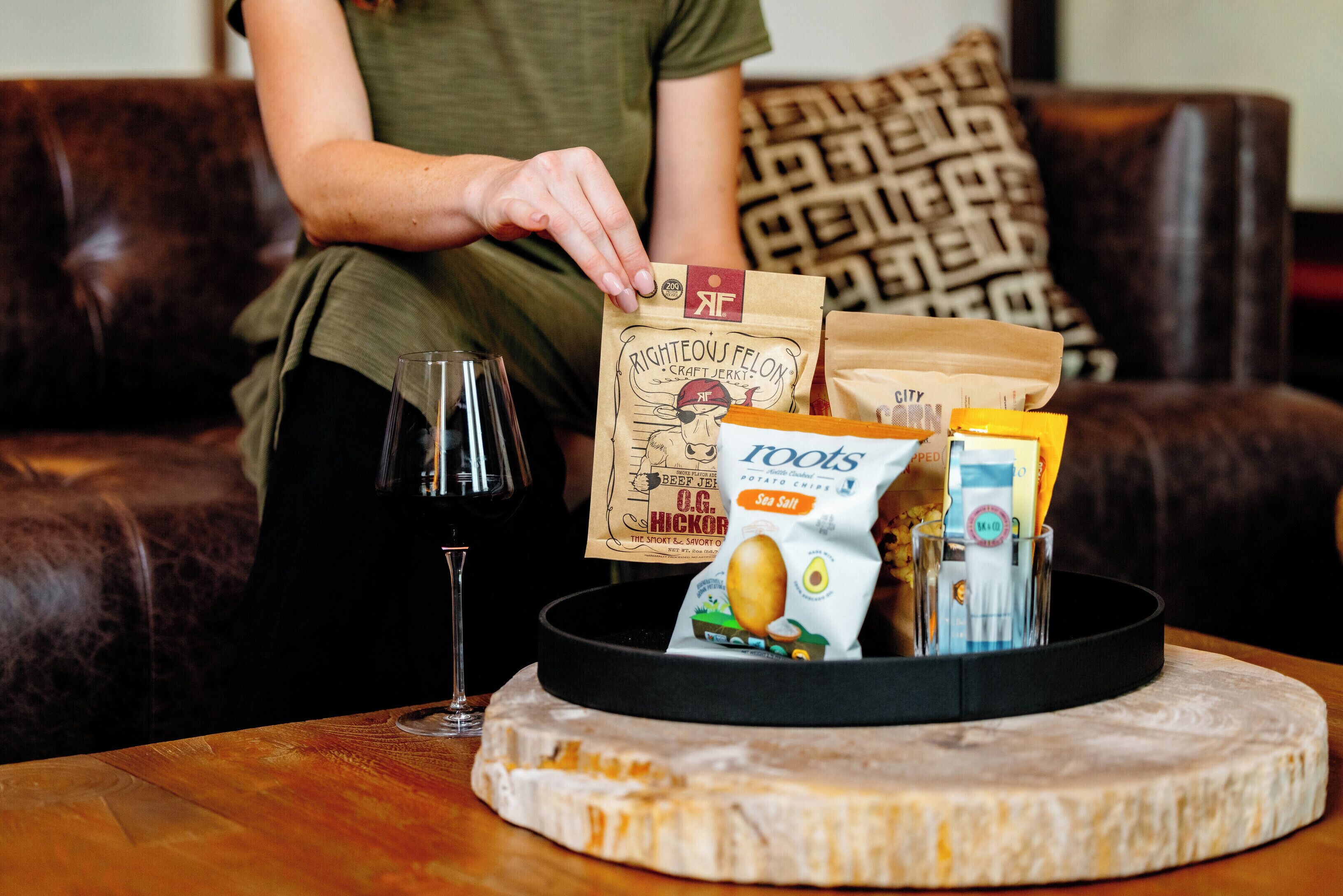 Una persona sentada en un sofá con una bandeja que contiene un queso grande, una copa de vino tinto y diversas botanas empaquetadas.