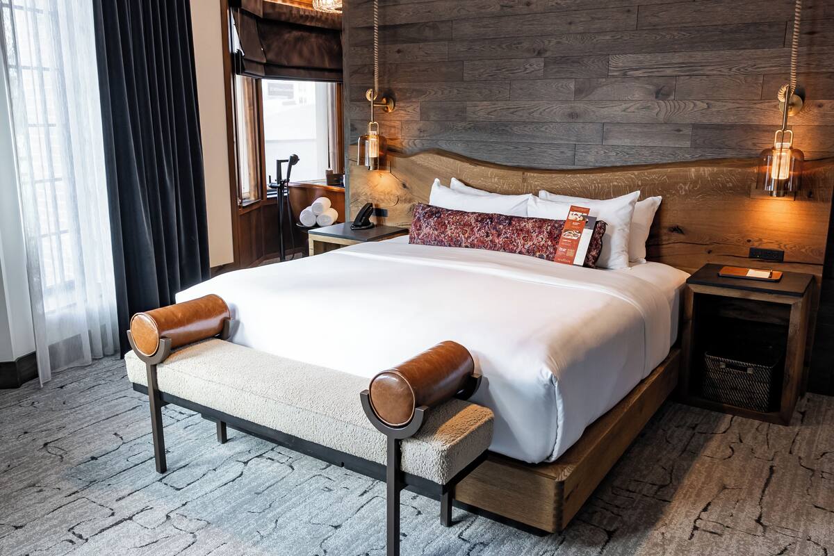 A hotel room with a wooden headboard, a large bed with white linens, a bench with leather cushions, a bedside table with a lamp, and a window with curtains.