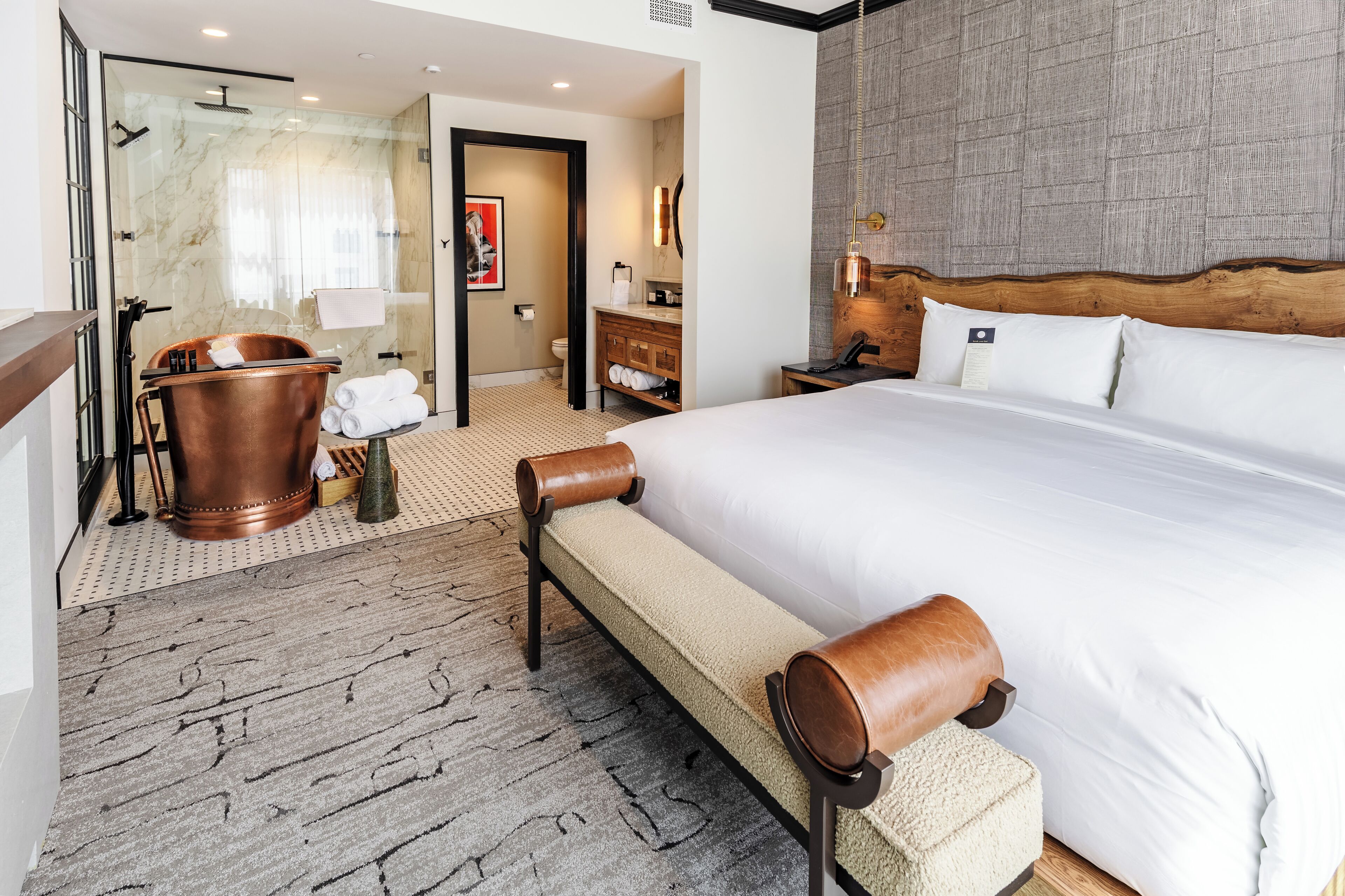 A hotel room with a large bed, a wooden headboard, a glass shower enclosure, a wooden bench, and a small table with a lamp.
