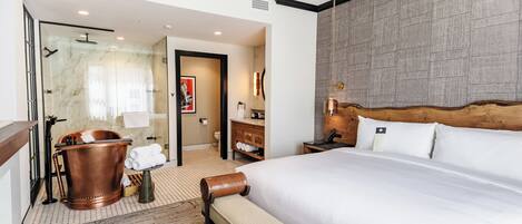 A hotel room with a large bed, a wooden headboard, a glass shower enclosure, a wooden bench, and a small table with a lamp.