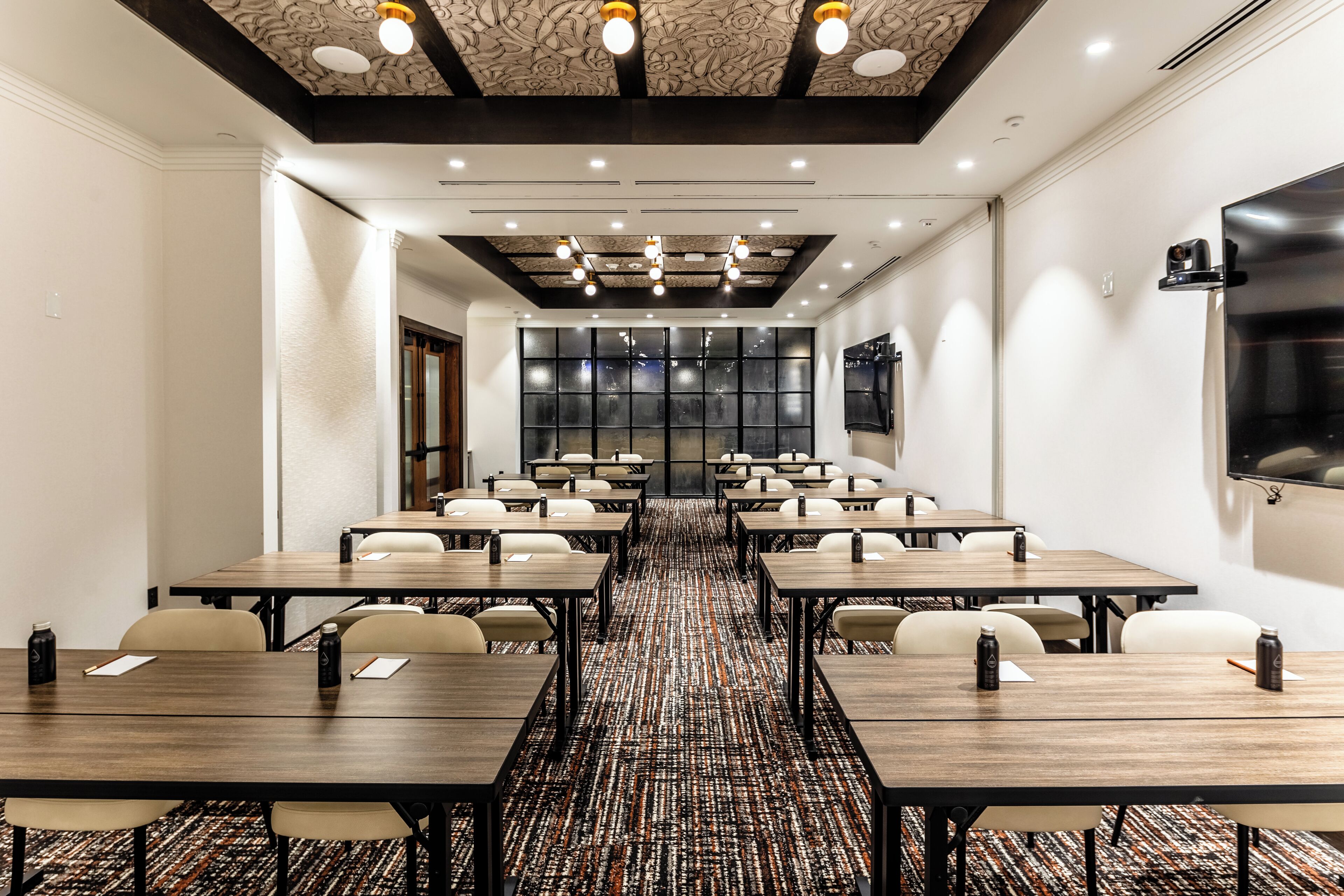 Un salón de conferencias con varias mesas y sillas, alfombra estampada y un diseño moderno de techo.
