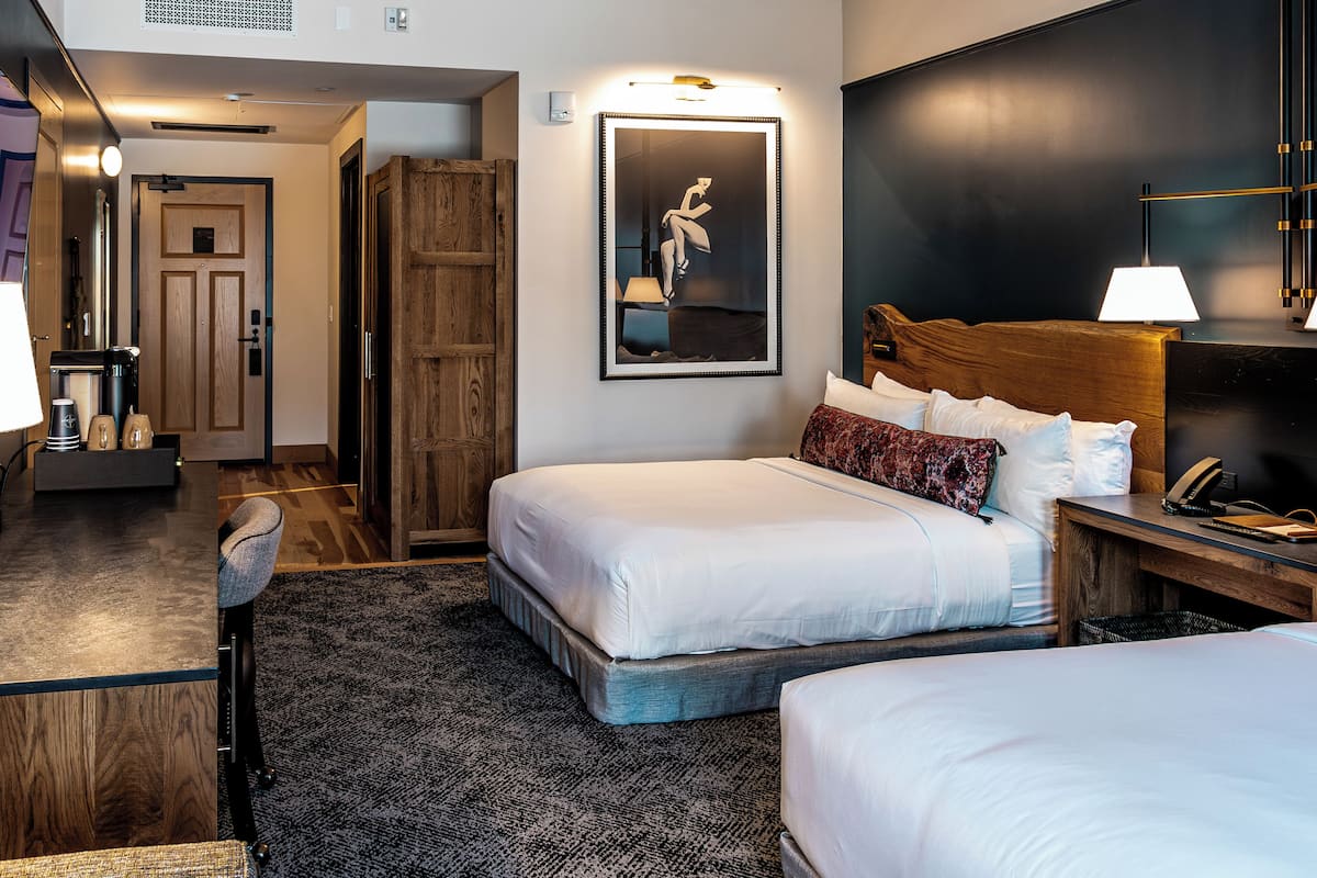A hotel room with two beds, a wooden wardrobe, a desk with a lamp, and a framed picture on the wall.